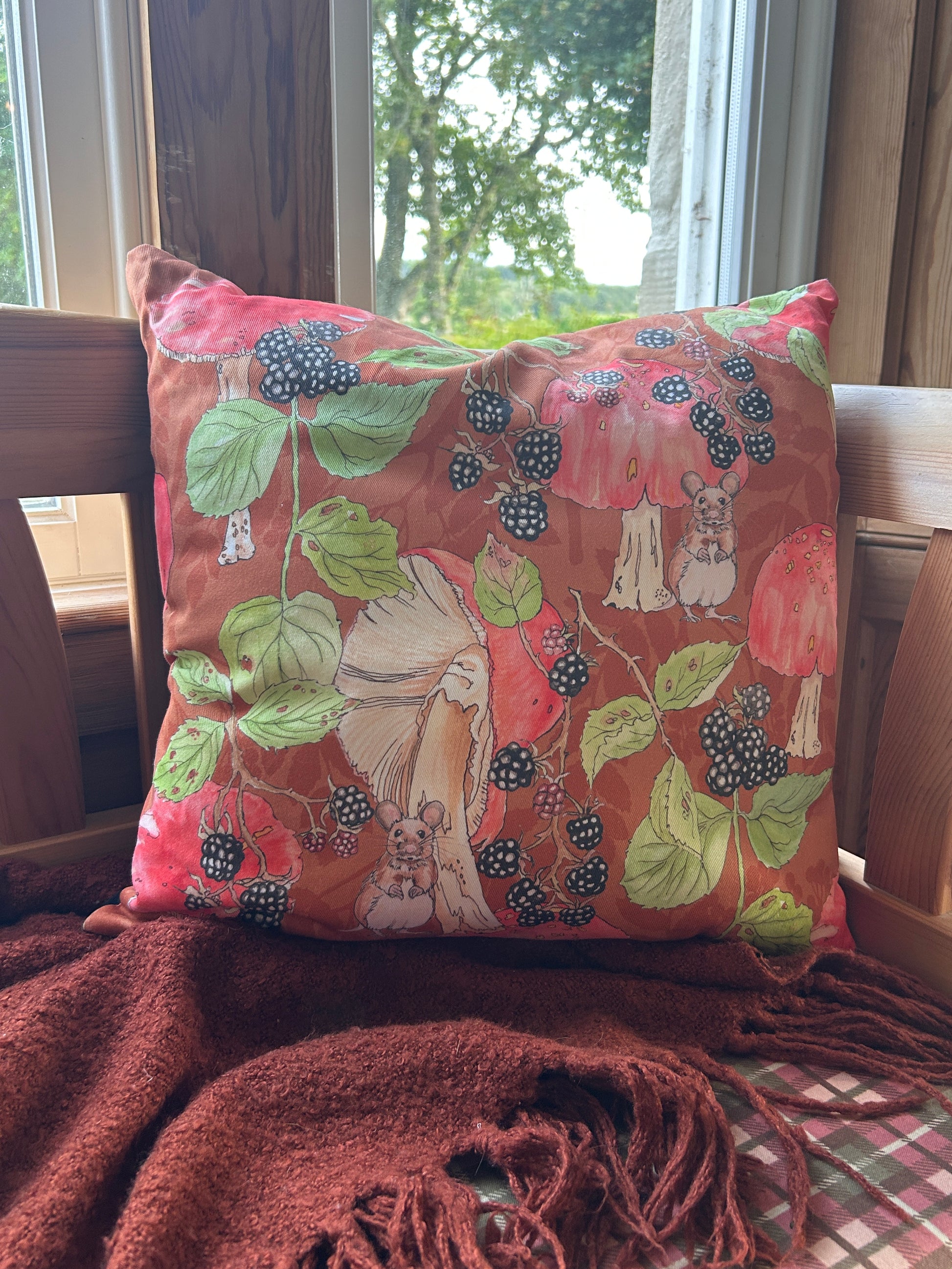 A decorative cushion in a blackberries, mushrooms, mice print with rust background. Resting on a wooden chair with a brown fringed blanket.