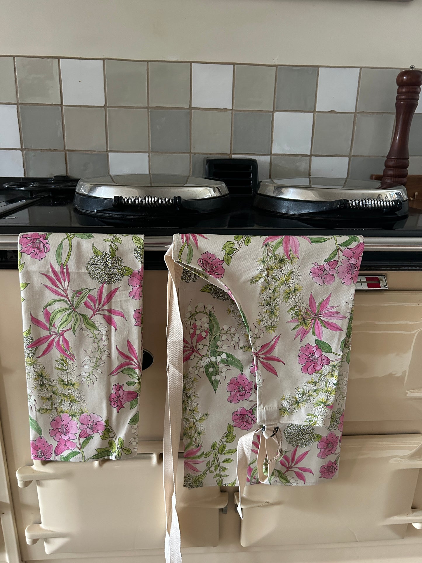 Floral-patterned tea towel and matching apron hanging on a stove with tiled wall in the background
