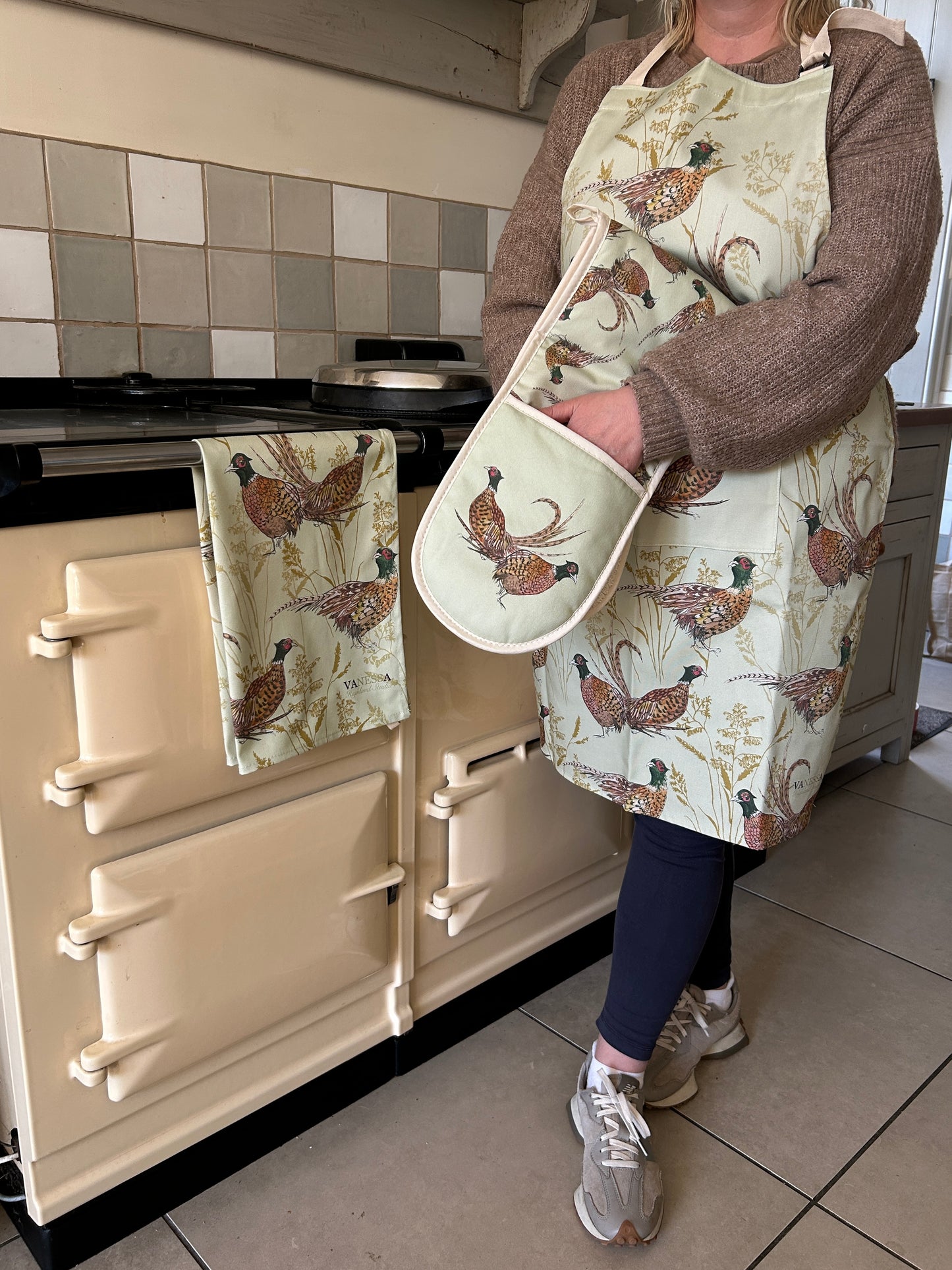 Person standing beside an AGA wearing an apron, oven glove and tea towel in a pheasant illustrated design on mint green background