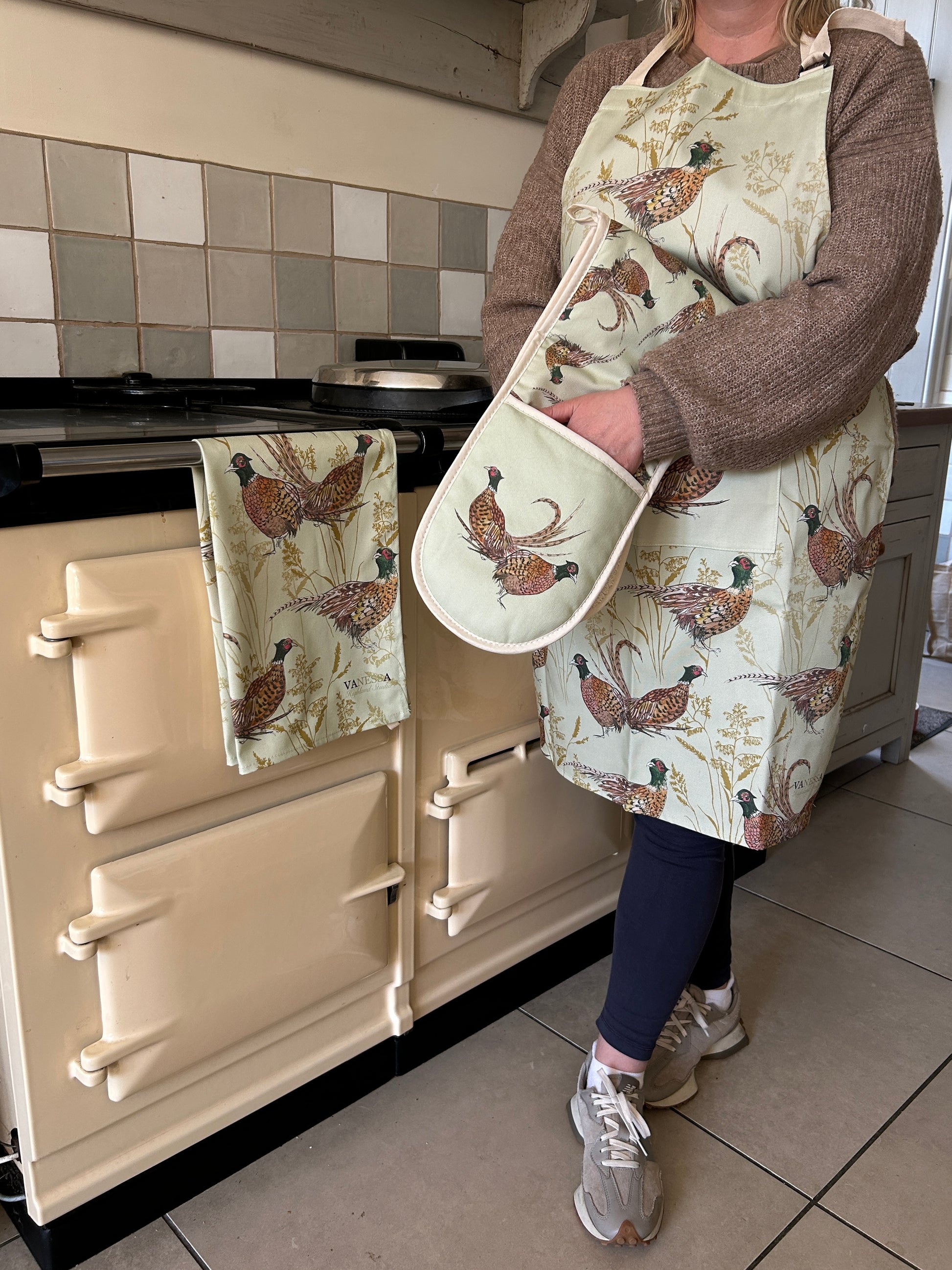 Person standing beside an AGA wearing an apron, oven glove and tea towel in a pheasant illustrated design on mint green background