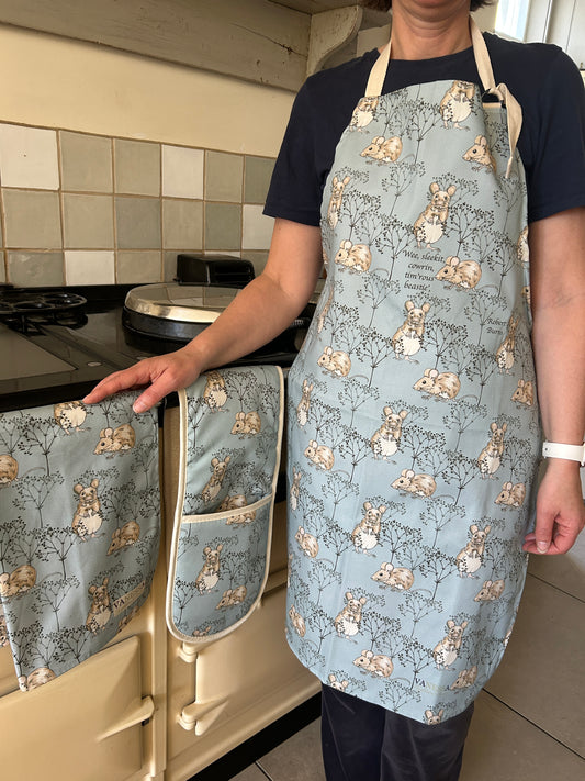 Person standing by range cooker with mouse design apron in Blue beside hanging oven gloves and tea towel