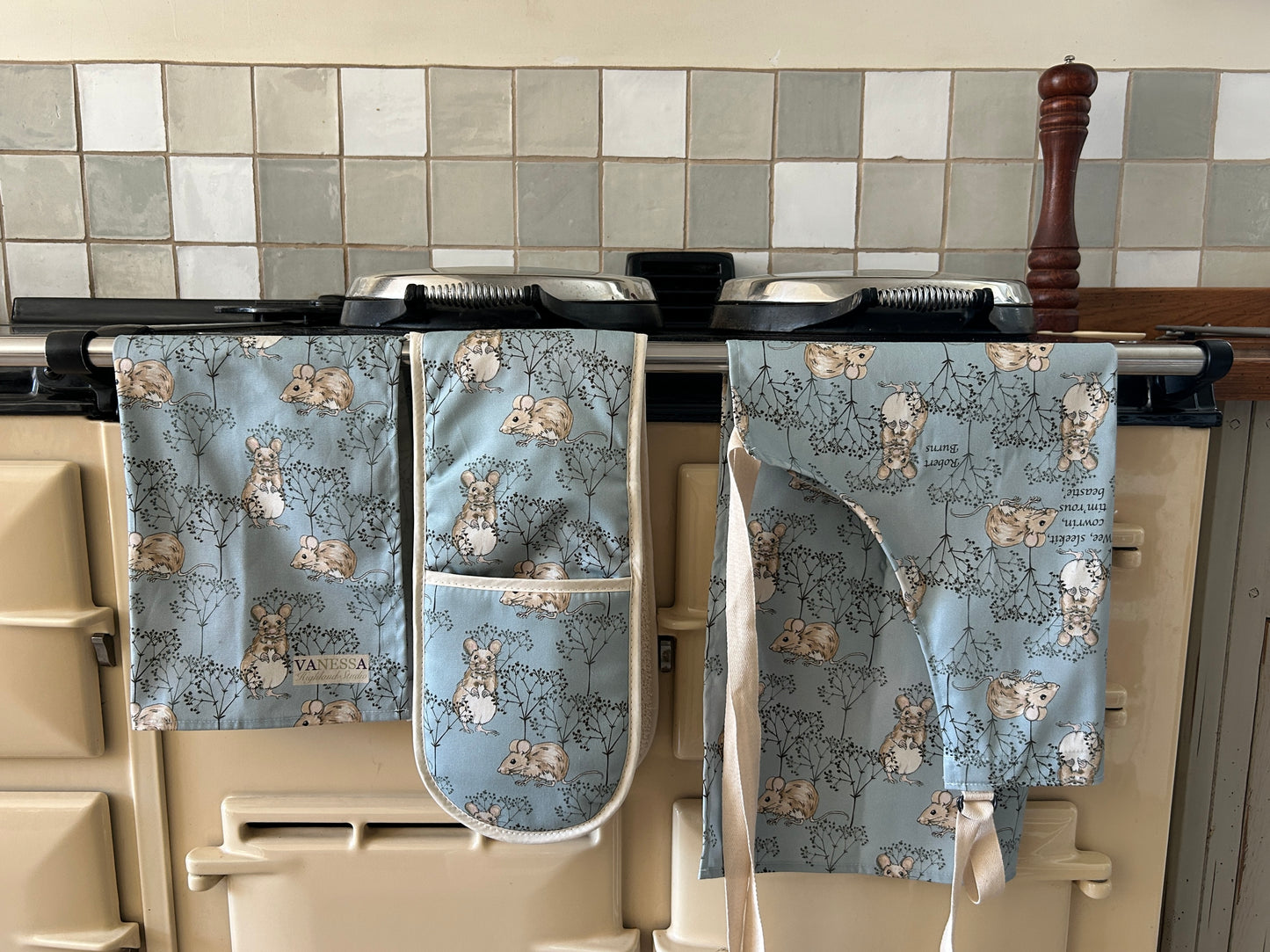 Set of kitchen apron, oven mitt, and tea towel with little mouse pattern in soft Blue, hanging on a stove.