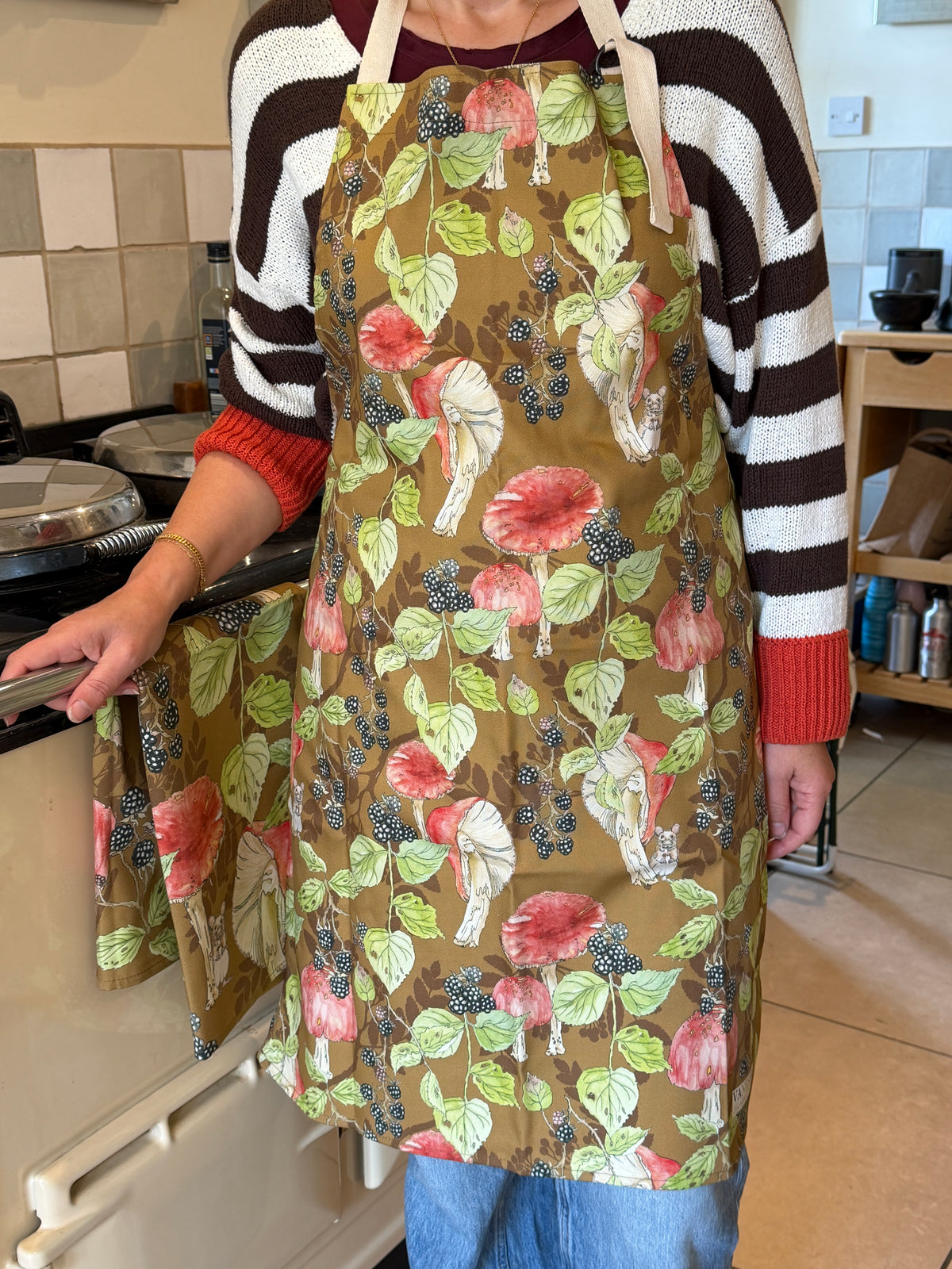 Person wearing an apron with toadstools and black berries design on tan back, standing beside a range cookerround