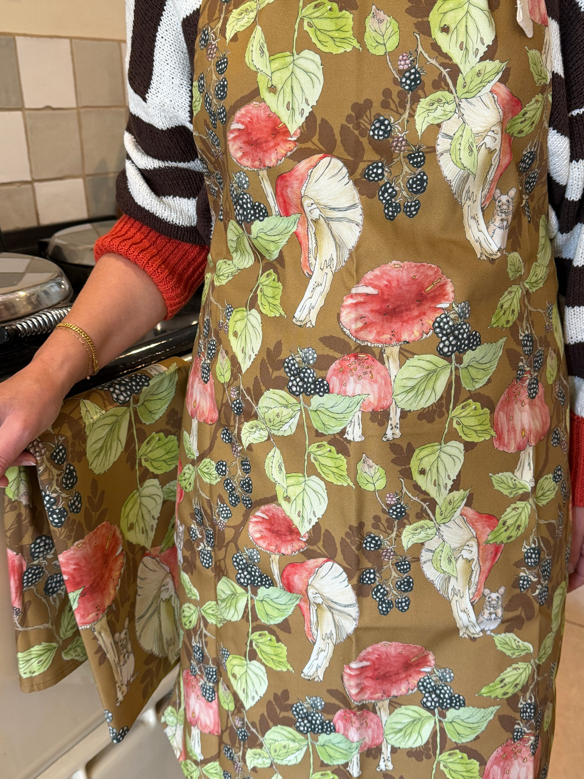 A person wearing a brown apron with a colorful mushroom and berry print, standing in a kitchen next to a range cooker. The apron features hand painted toadstools and black berries with a brown background.