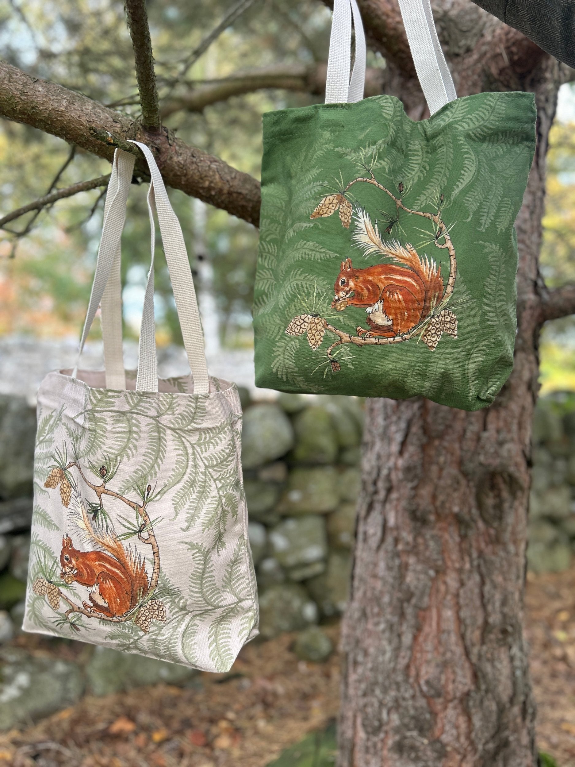 Red Squirrel and fern tote bags. cream design background, cream design background. Both hanging on a pine branch