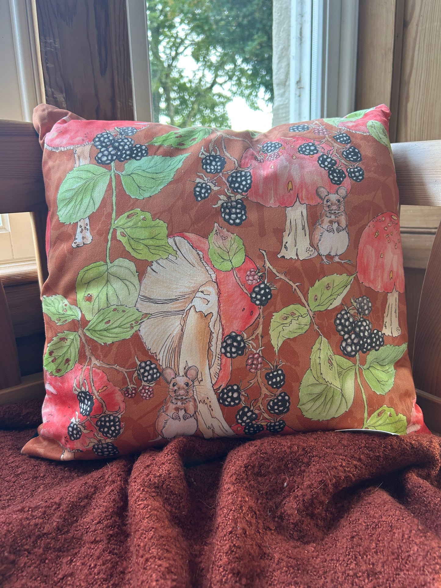 A cushion with a woodland print of mushrooms, Brambles and mice on a rust-orange background. Featured sitting on a throw in the window