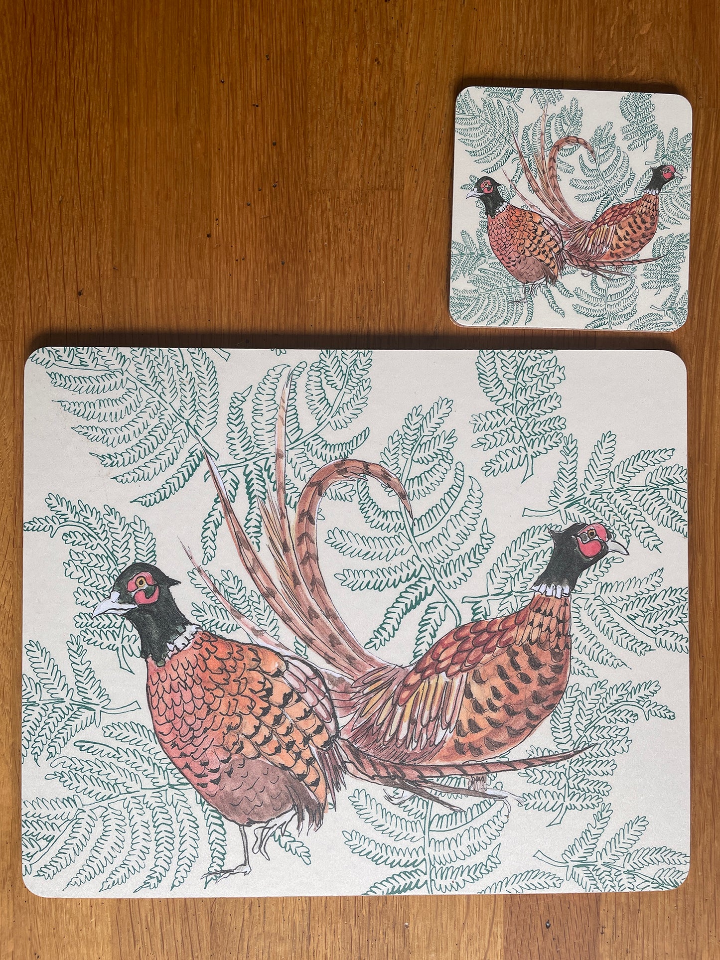 Matching placemat and coaster featuring pair of pheasants with ferns. Beige background.