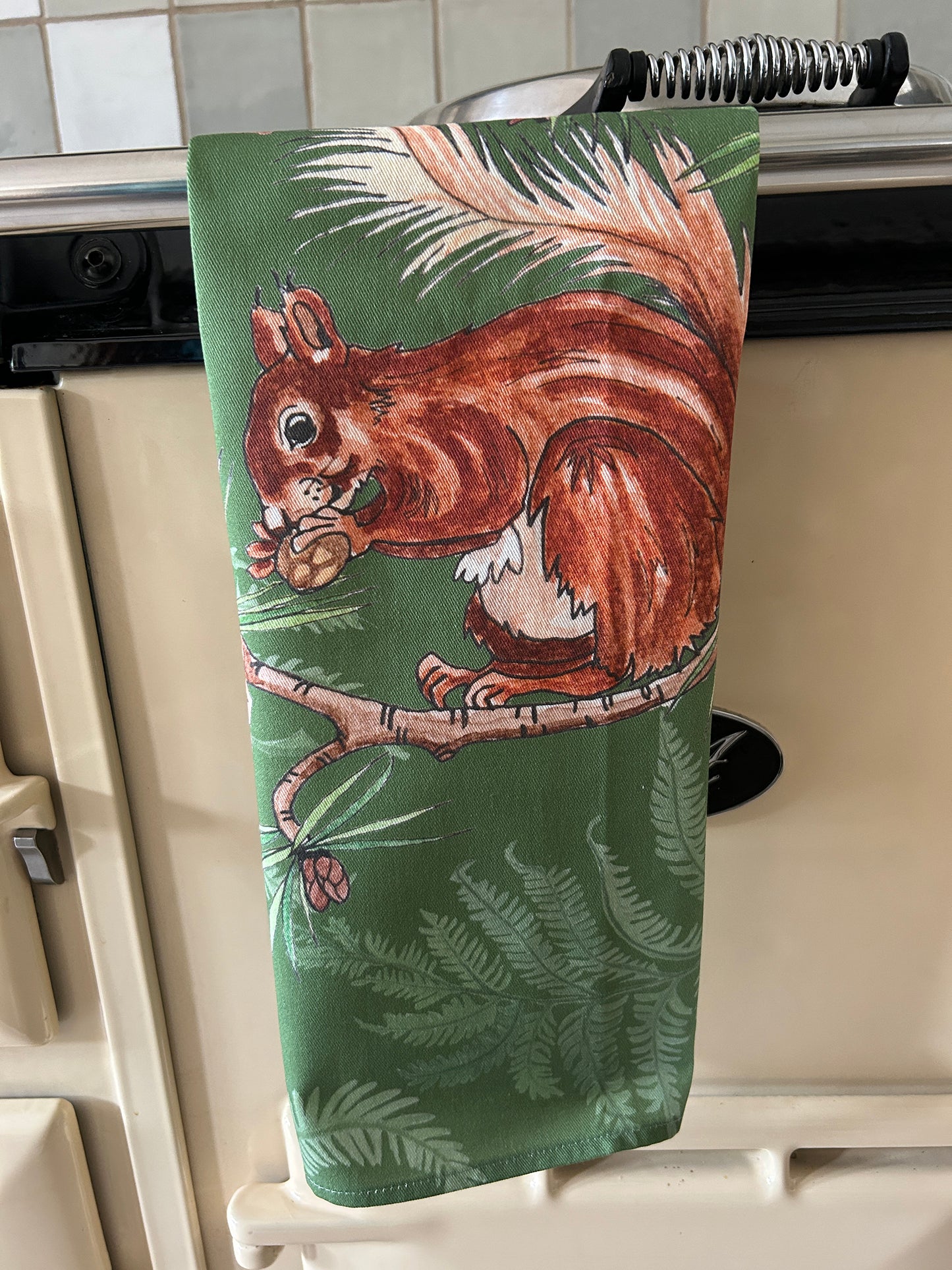 Single red squirrel design with fern background in green, printed on cotton tea towel hanging on Aga